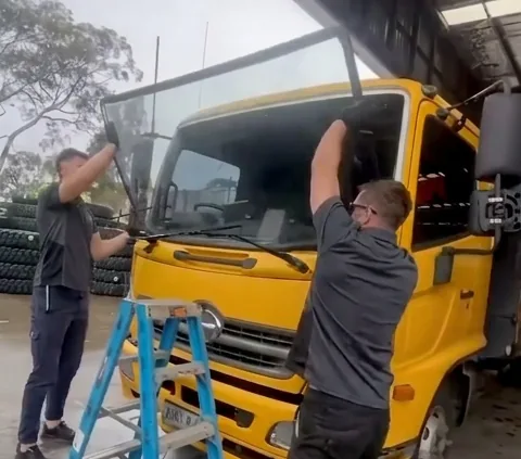 Two technicians lifting a windscreen into the cab of a yellow truck, one working from a stepladder.