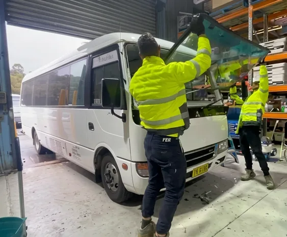 Two technicians lifting a large windscreen into place on a minibus, with a second pair working behind them.