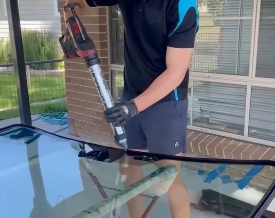 A technician applying adhesive to a windscreen under cover on the driveway of a house.