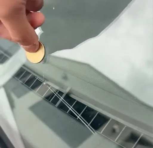 A hand holding a coin flat against a windscreen beside a crack, using it to judge whether the damage is small enough to repair.